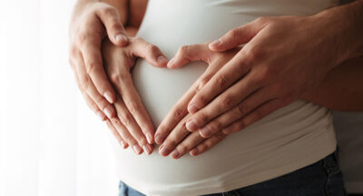 Close up portrait of man's and woman's hands making heart gesture over pregnant belly indoors